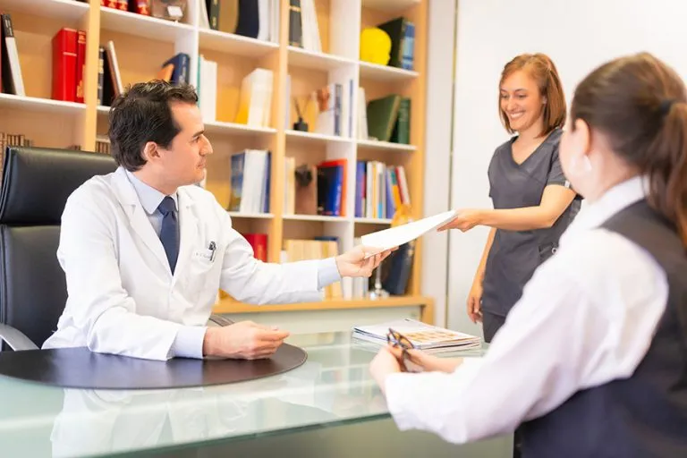 Dr. César Casado Sánchez, cirujano plástico experto en tratamientos faciales y corporales, posa sonriente y profesional en su consulta, destacando su experiencia.